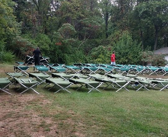 A field of green grass covered in cots. People are moving and cleaning the cots. They are preparing the cots for Winter Relief. 